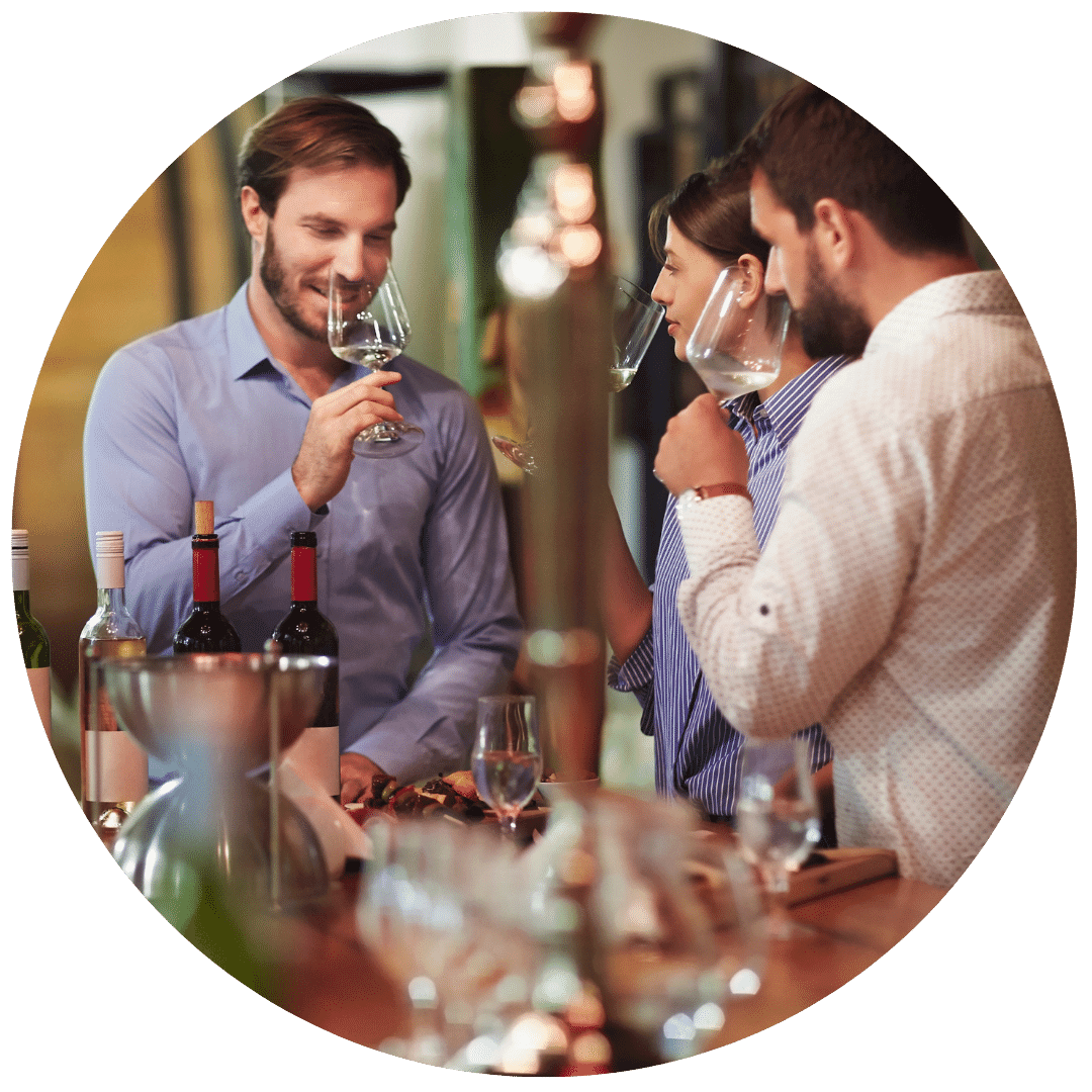 two men and one woman on a wine tasting tour, sniffing their wine glasses with various bottles of wine on the table next to them