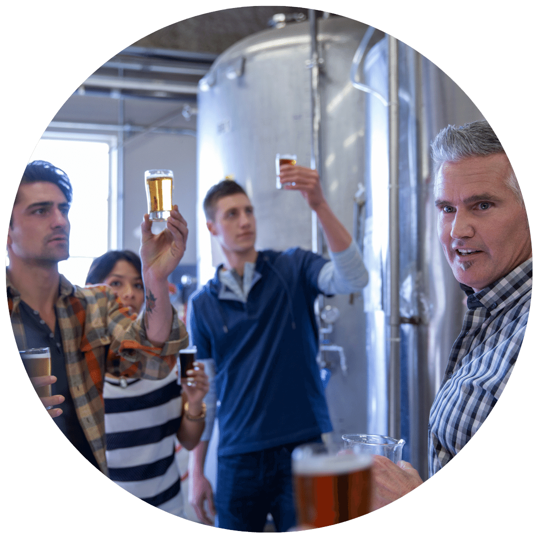 middle aged man with salt and pepper hair in a button-down shirt leading a tour of a brewery with three young people in the background holding up various glasses of beer